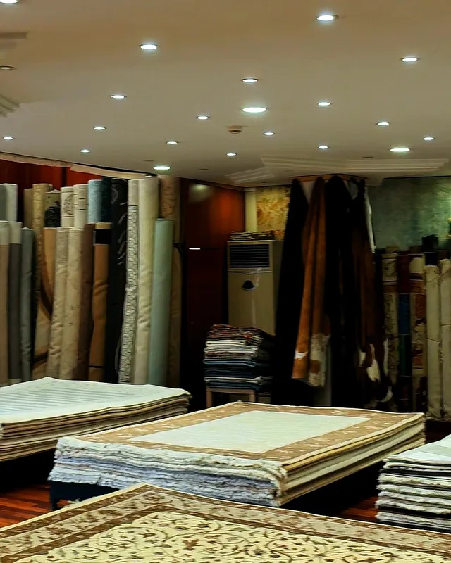 Mağazamızın içi: ahşap zeminde üst üste serili halı yığınları, arkada duvar boyunca dizili rulo halılar