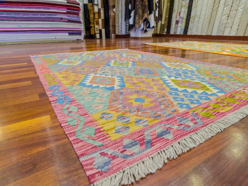 Mağazamızın ahşap zemininde pembe bordürlü, mavi, hardal ve yeşil baklava desenli pastel kilim; arkada katlı halılar ve rulo halılar