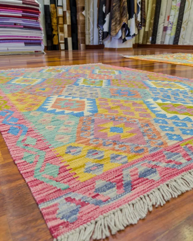 Mağazamızın ahşap zemininde pembe bordürlü, mavi, hardal ve yeşil baklava desenli pastel kilim; arkada katlı halılar ve rulo halılar