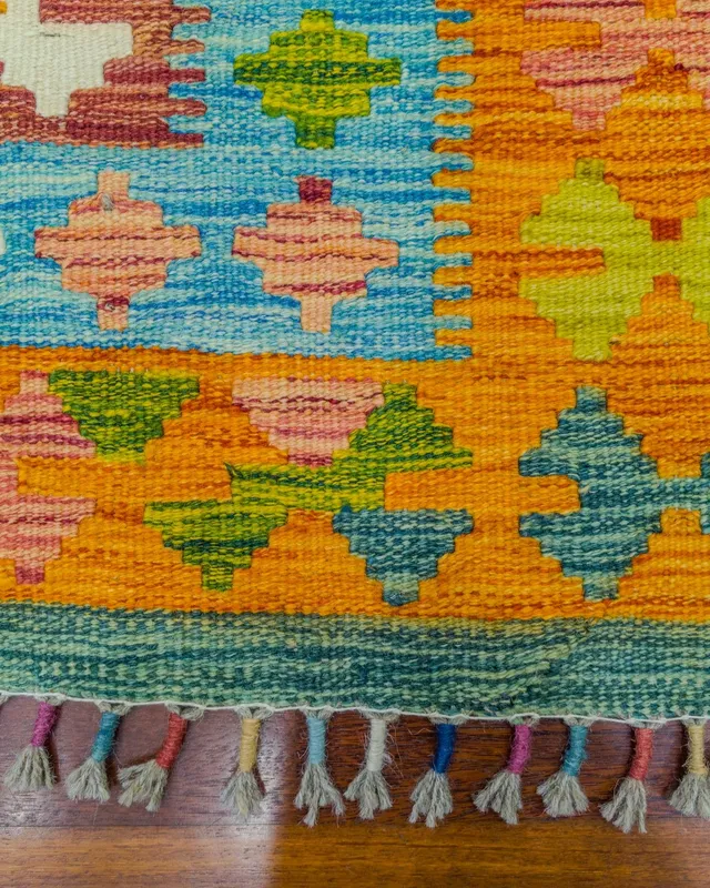 Kilim köşesinin yakın planı: turuncu bordür, renk geçişleri ve elle sarılmış çok renkli püskül uçları